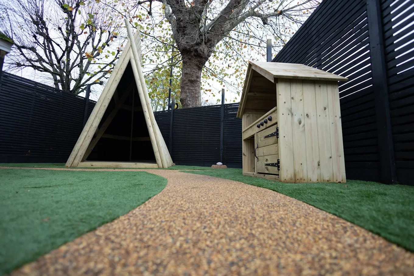 Timber play shelters beside the dark garden fence at Ealing